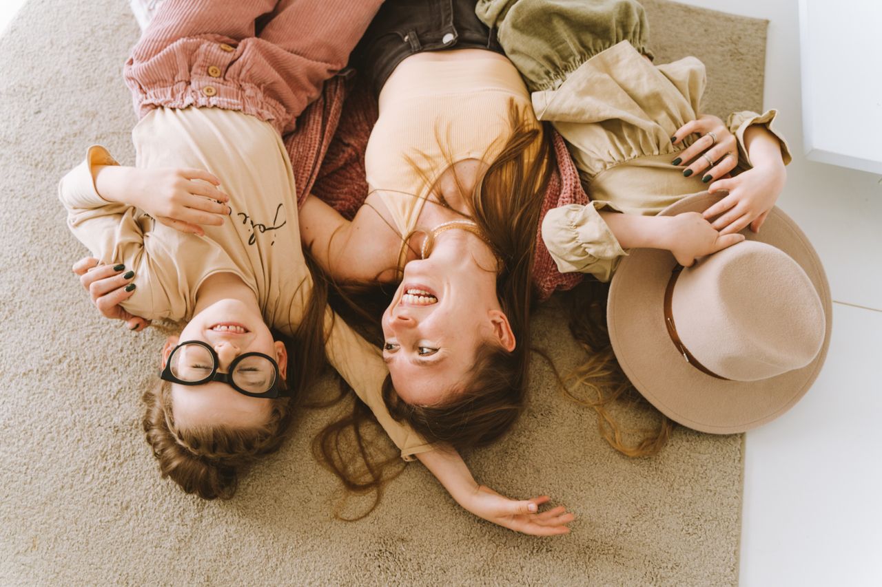 founder-img Mother and daughters lying happily on the carpet, embracing and smiling.